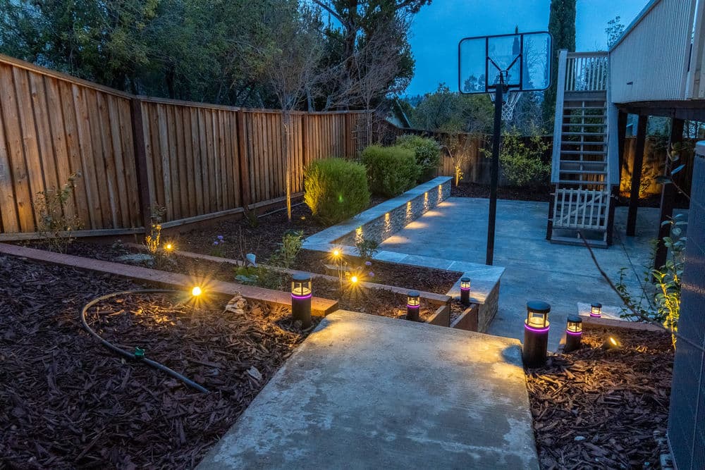 Modern backyard landscape with ambient lighting, basketball hoop, and slate seating area.
